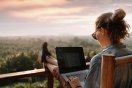 Person som jobber på laptop med utsikt over skog og solnedgang.