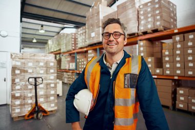 Mannlig arbeidsleder innen logistikk står med vernehjelm under armen i et varelager og smiler