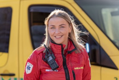 Ambulansearbeider i rød uniform foran en gul ambulanse, smilende og klar til innsats.