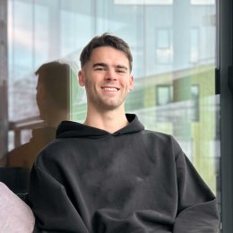 Smilende mann i svart hettegenser sitter foran et vindu.