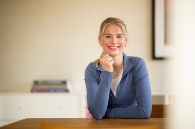 Smilende kvinne i blå genser sitter ved et bord med bøker i bakgrunnen.