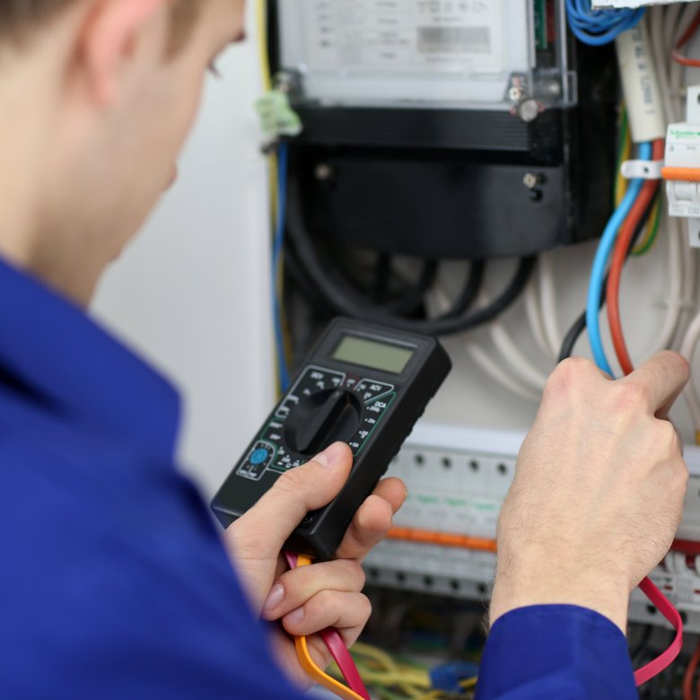 Elektriker i blå uniform bruker multimeter for å måle spenning i et sikringsskap.