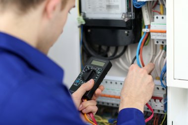 Elektriker i blå uniform bruker multimeter for å måle spenning i et sikringsskap.