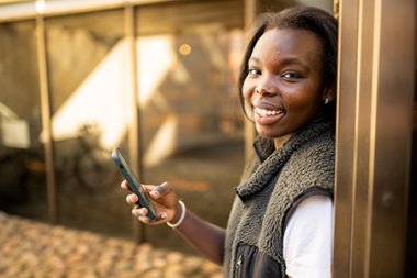 Person smiler og holder en mobiltelefon, står lent mot en vegg utendørs.