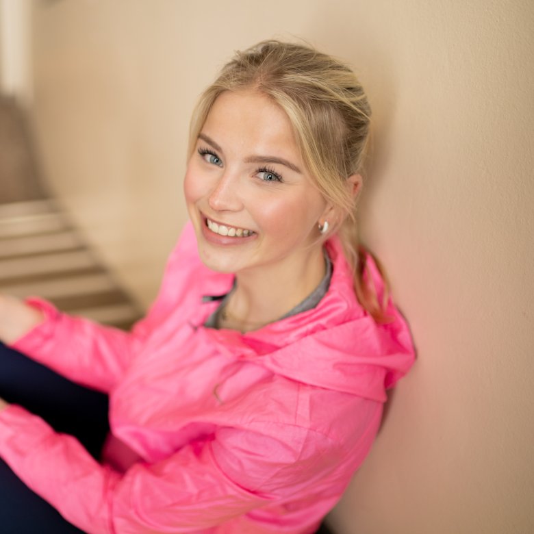 Ung kvinne i rosa jakke smiler og sitter på en trapp.