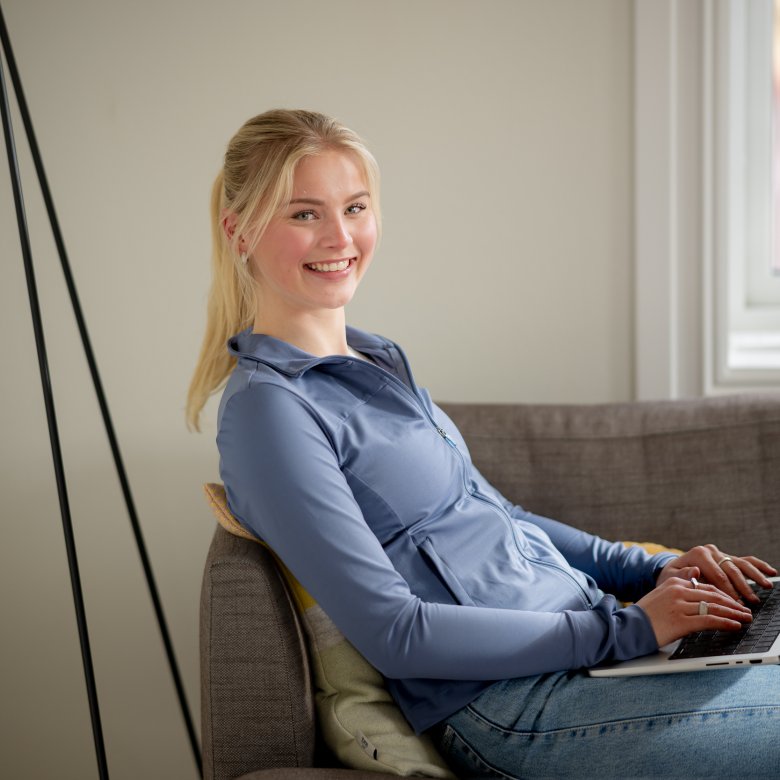 Kvinne smiler mens hun jobber på en bærbar datamaskin i en sofa.