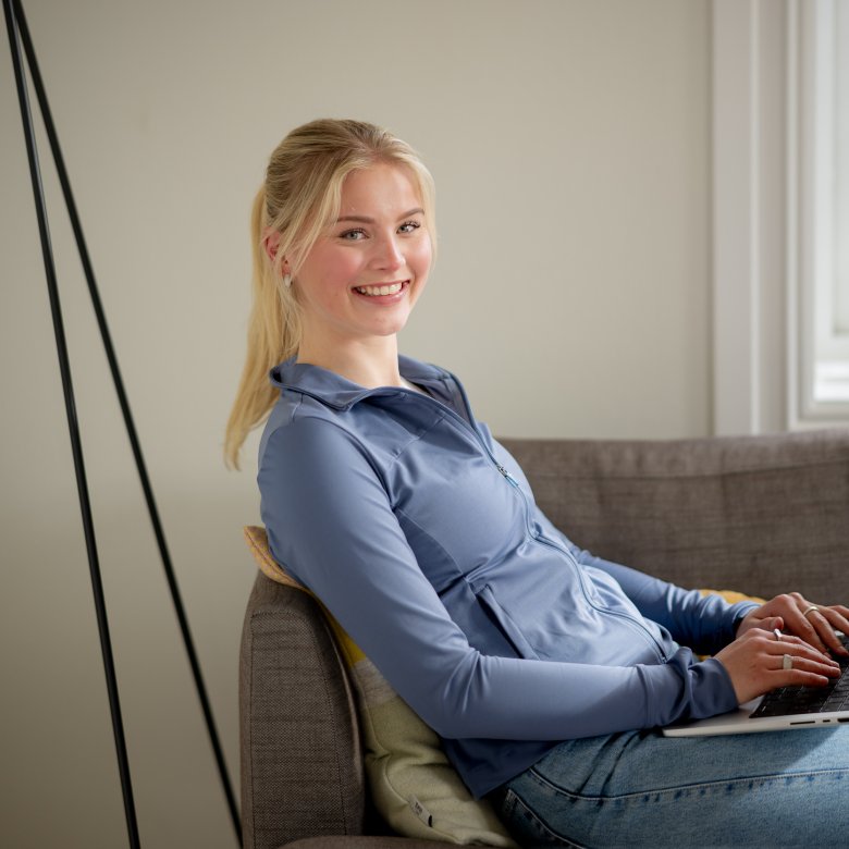 Kvinne smiler mens hun jobber på en bærbar datamaskin i en sofa.