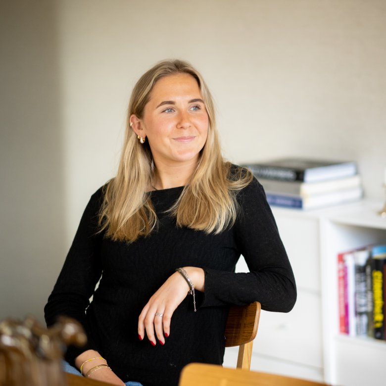 Kvinne med langt blondt hår sitter ved et bord og smiler.