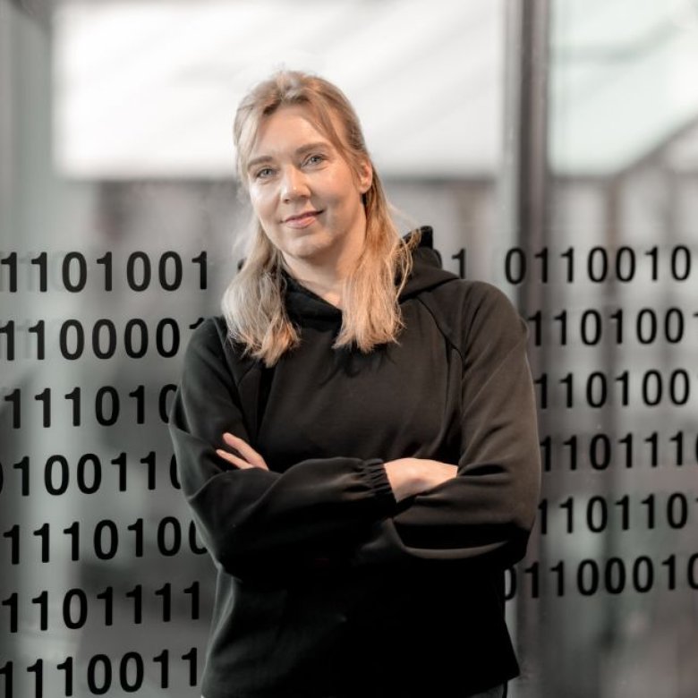 Kvinne står foran et vindu med binære tall, kledd i svart genser.