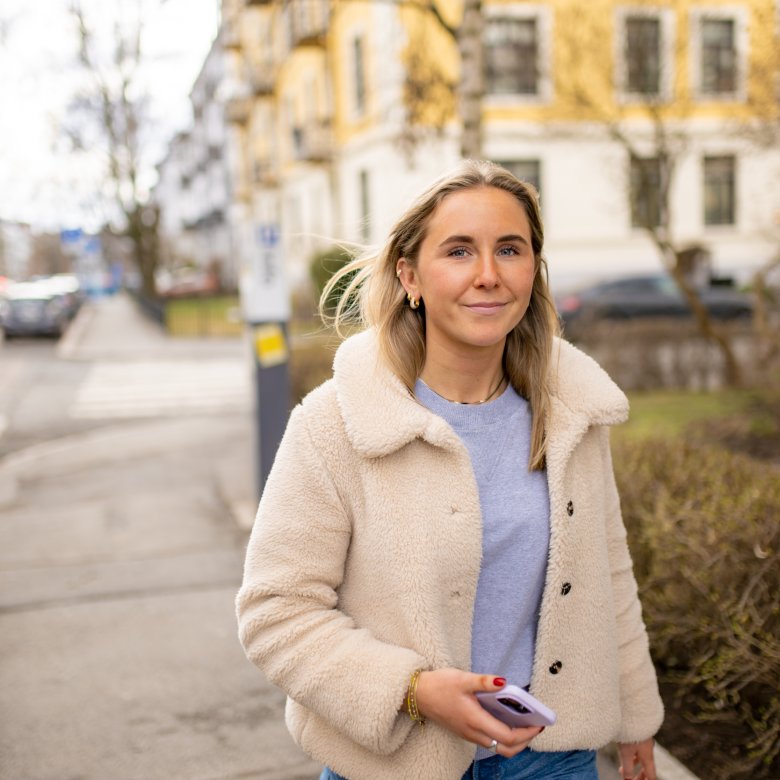 Kvinne med beige jakke og mobiltelefon går langs en gate med bygninger i bakgrunnen.