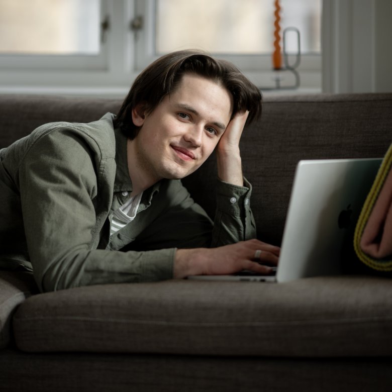 Ung mann smiler mens han jobber på en bærbar datamaskin i en sofa.