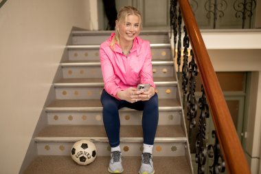 Kvinne i rosa jakke sitter på trapp med fotball ved siden av seg.