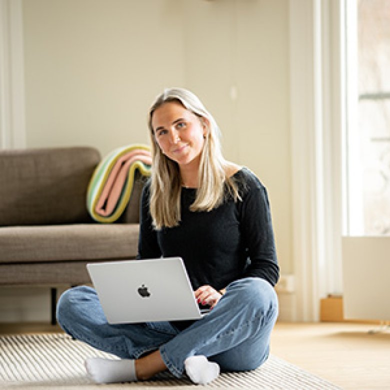 Kvinne sitter på gulvet med en bærbar datamaskin i et lyst rom.