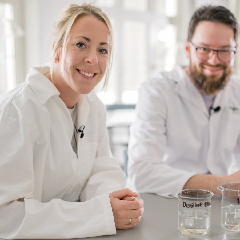 To personer i laboratoriefrakker smiler ved et bord med begerglass.