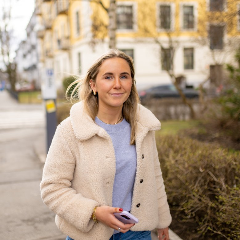 Kvinne i lys jakke går utendørs med en mobiltelefon i hånden.