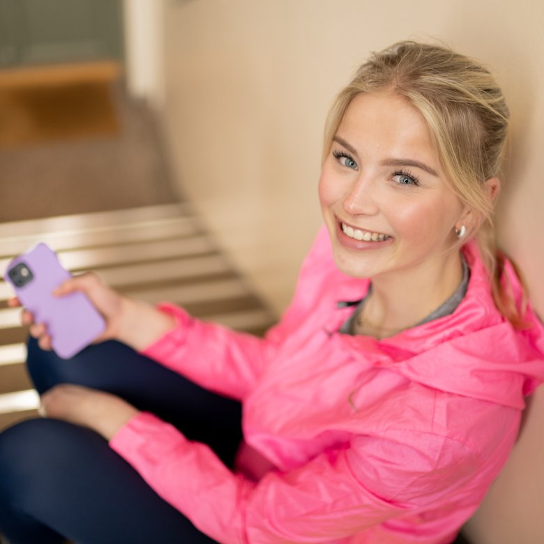 Ung kvinne i rosa jakke smiler og holder en mobiltelefon.