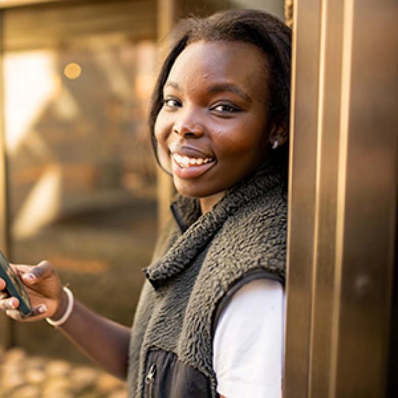 Person smiler og holder en mobiltelefon, står lent mot en vegg utendørs.