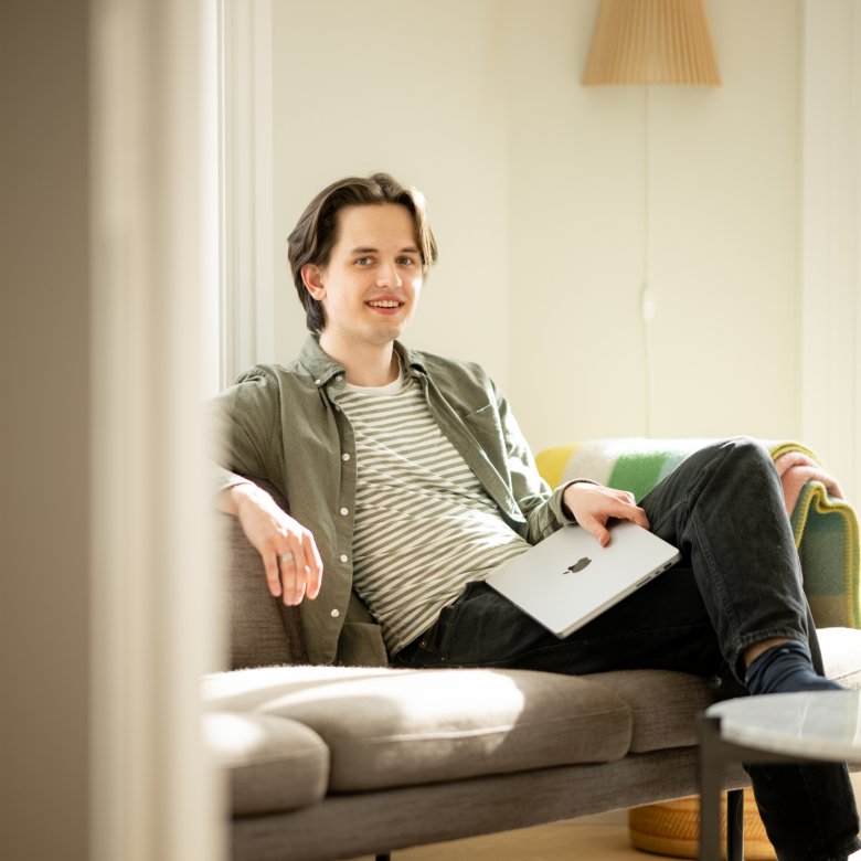 Person sitter på sofa med laptop i solfylt rom.