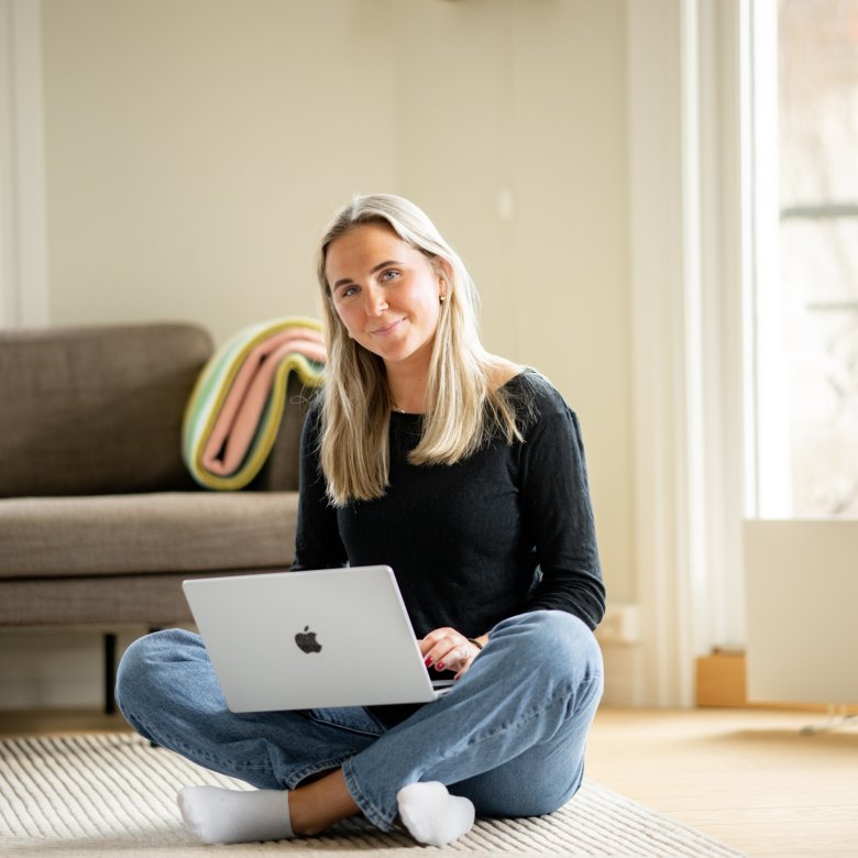 Kvinne sitter på gulvet med en laptop i et lyst rom.