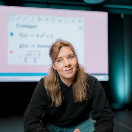 Kvinne foran en skjerm med matematiske formler, smilende og iført svart genser.