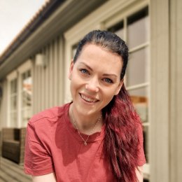 Kvinne i rød t-skjorte smiler foran et hus med store vinduer.