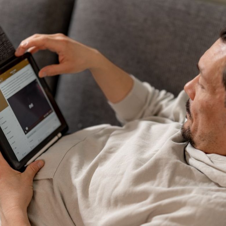 Mann med hodetelefoner ser på nettbrett, liggende på sofa.