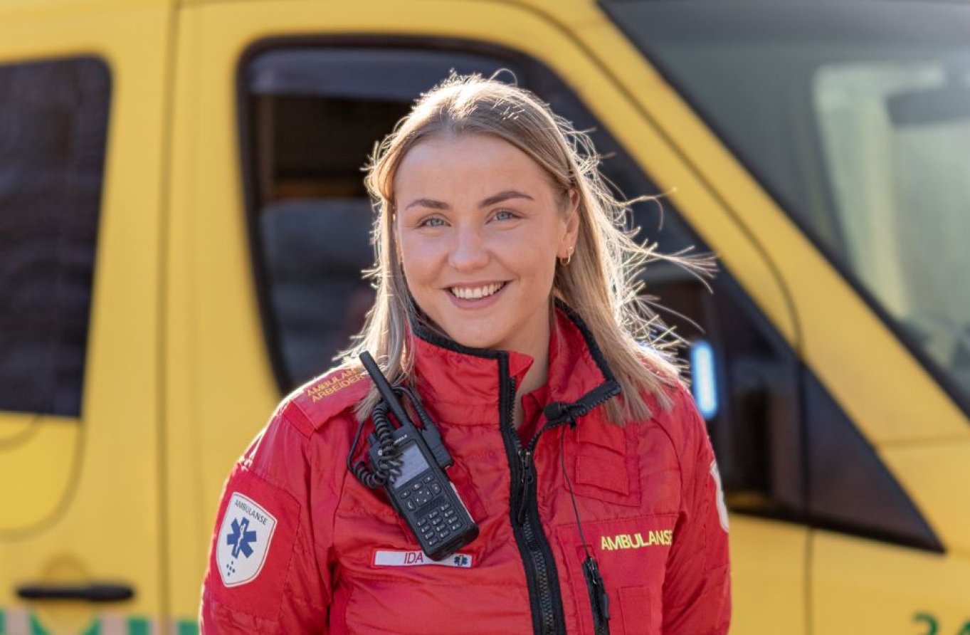 Ambulansearbeider i rød uniform foran en gul ambulanse, smilende og klar til innsats.