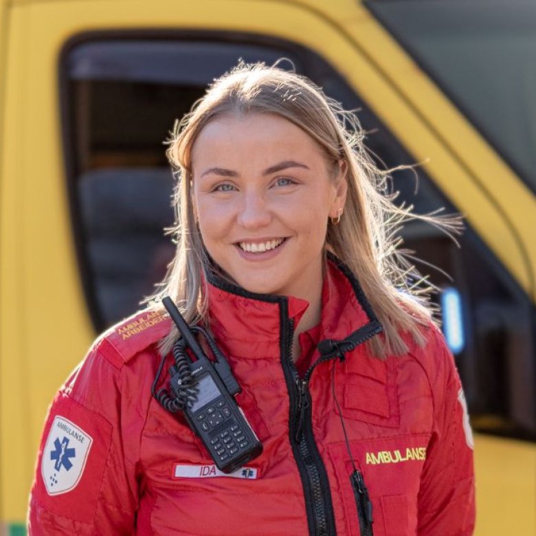 Ambulansearbeider i rød uniform foran en gul ambulanse, smilende og klar til innsats.