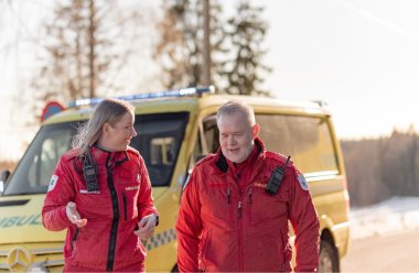 To ambulansearbeidere i røde uniformer står foran en gul ambulanse i solskinn.