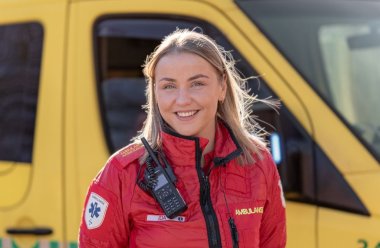 Ambulansearbeider i rød uniform foran en gul ambulanse, smilende og klar til innsats.