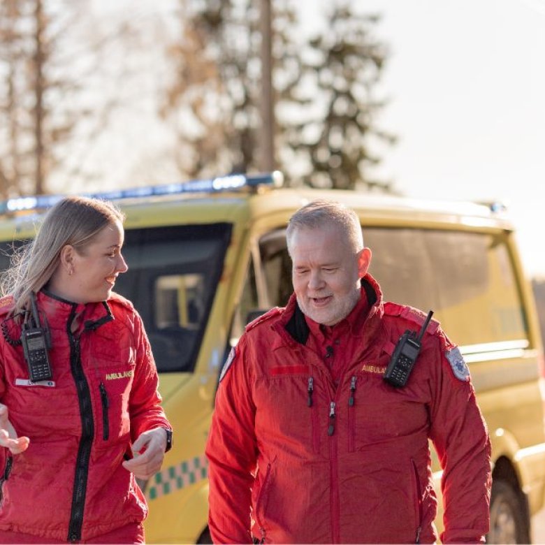 To ambulansearbeidere i røde uniformer står foran en gul ambulanse i solskinn.